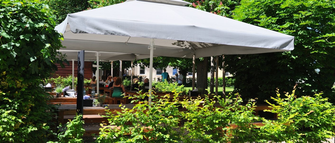 A large parasol covers seats in a garden restaurant surrounded by lush greenery and trees., © Schlosshotel Monrepos A large parasol covers seats in a garden restaurant surrounded by lush greenery and trees., © Schlosshotel Monrepos