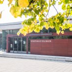 The Stadthalle Eislingen with its modern glass façade and red entrance area, surrounded by autumn leaves., © GIACINTO_CARLUCCI