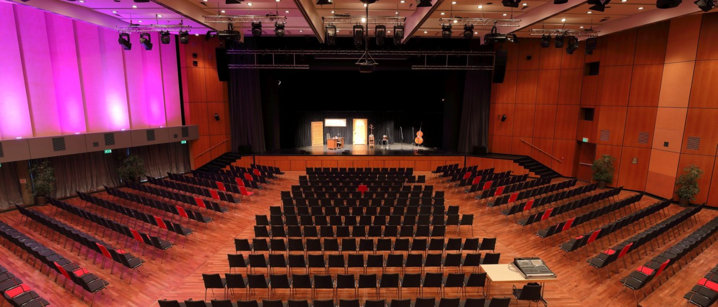 Large event hall with stage, empty rows of chairs and pink illuminated walls. The stage is prepared for a performance., © ccbs_gf Large event hall with stage, empty rows of chairs and pink illuminated walls. The stage is prepared for a performance., © ccbs_gf