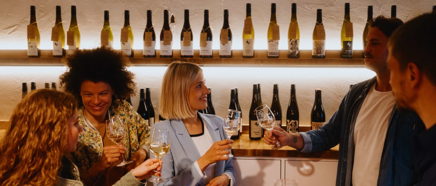 Menschen in einer Vinothek stoßen mit Weingläsern an. Im Hintergrund sind Weinflaschen auf Regalen zu sehen., © SMG, Alwin Maigler