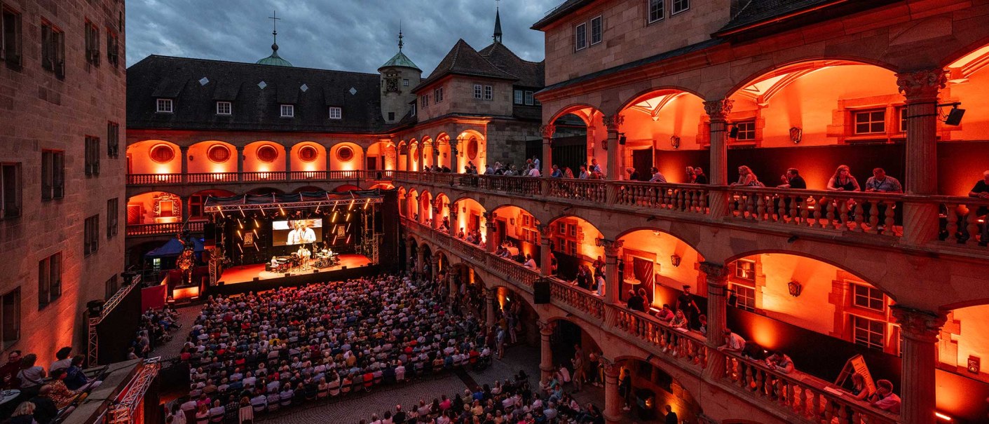 Open-air concert in the courtyard of a historic building, illuminated in red. Spectators sit on balconies and in the courtyard, stage with musicians., © jazzopen stuttgart, Reiner Pfisterer
