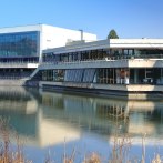 The Böblingen Congress Hall is reflected in the water, surrounded by bare trees and reeds, under a clear blue sky., © ccbs_gf
