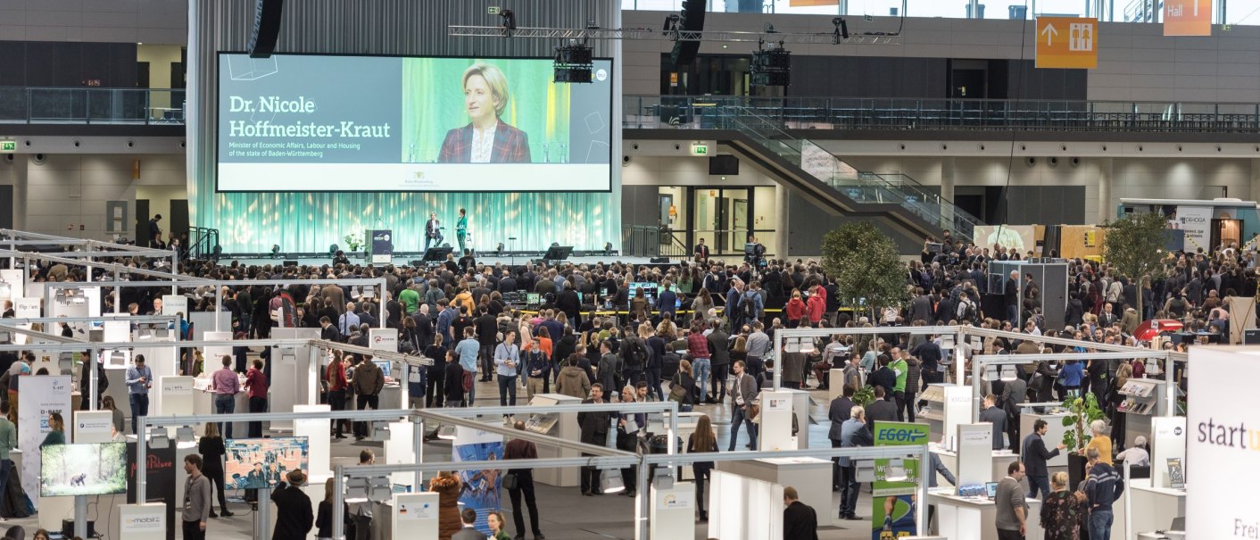 Large exhibition hall in Stuttgart with many visitors, stands and a stage with a large screen. Presentation by Dr. Nicole Hoffmeister-Kraut., &copy; Landesmesse Stuttgart GmbH / Valentin Marquardt