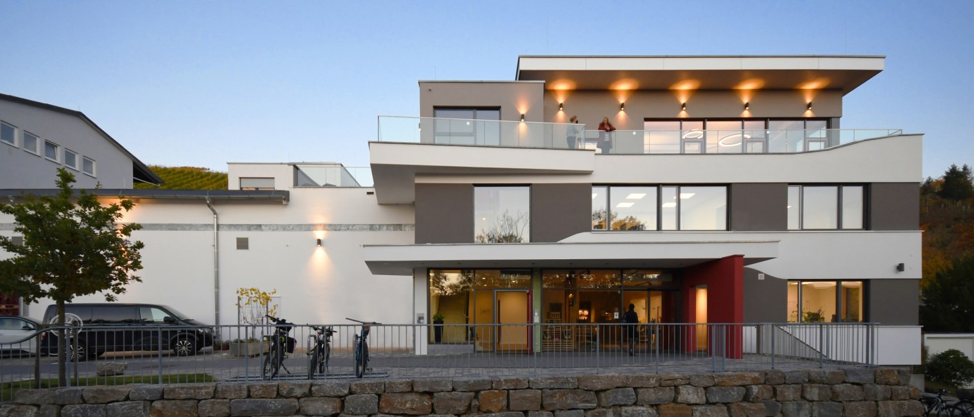 Moderne Gebäudeaußenansicht mit Glasbalkonen, Beleuchtung und Fahrrädern im Vordergrund. Abendstimmung mit klarer, blauer Himmel., © Architekurbuero-Gaysert