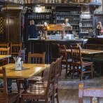 Gemütlicher Innenraum eines Restaurants mit Holztischen und -stühlen, einer Bar im Hintergrund und einem großen Kachelofen links., © SMG, Sarah Schmid
