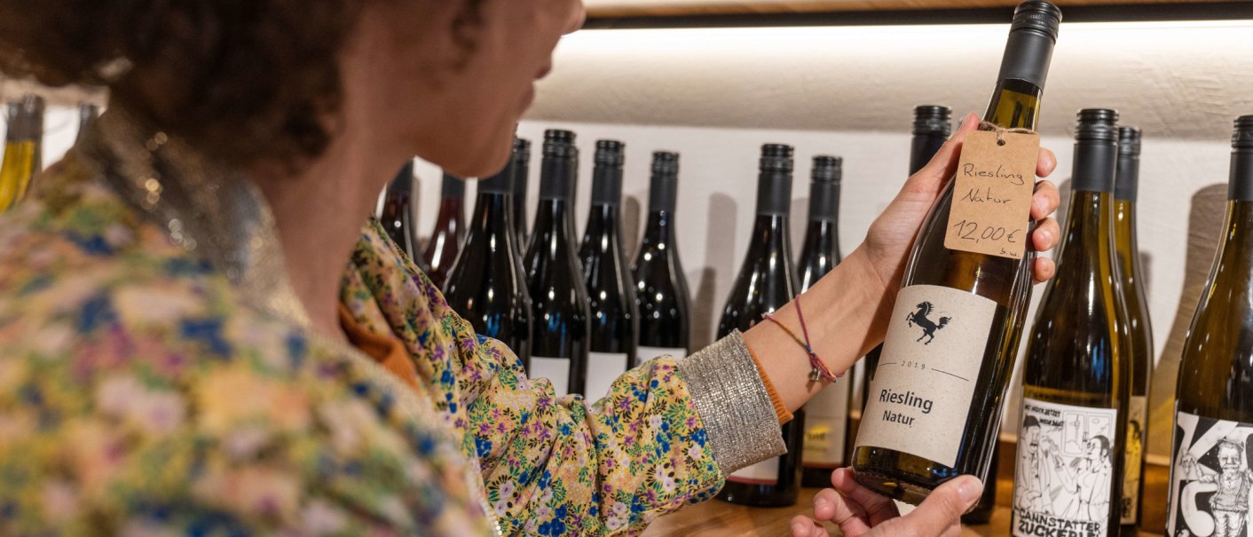 Eine Person betrachtet eine Weinflasche mit der Aufschrift 'Riesling Natur' in einer Weinbar. Im Hintergrund stehen weitere Weinflaschen., © SMG, Martina Denker