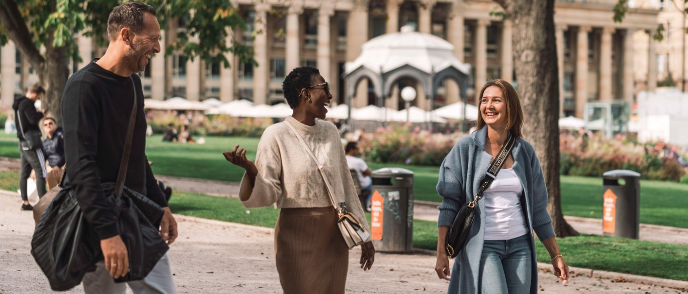 Drei Personen spazieren lachend im Oberen Schlossgarten. Im Hintergrund sind Bäume, ein Pavillon und ein Gebäude mit Säulen zu sehen., © SMG, wpsteinheisser Drei Personen spazieren lachend im Oberen Schlossgarten. Im Hintergrund sind Bäume, ein Pavillon und ein Gebäude mit Säulen zu sehen., © SMG, wpsteinheisser