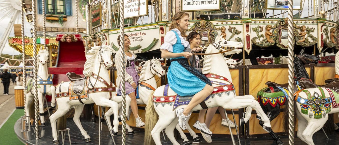 Zwei Frauen in Dirndl fahren auf einem nostalgischen Karussell mit wei&szlig;en Pferden. Im Hintergrund sind ein Riesenrad und ein Pizza-Stand zu sehen., &copy; Stuttgart-Marketing GmbH, Sarah Schmid