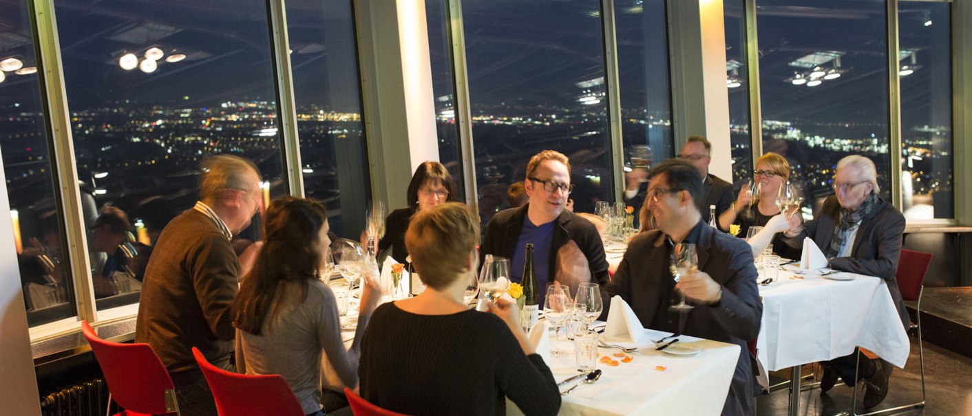 Gruppe von Menschen bei einem Abendessen in einem Restaurant mit Panoramablick auf eine beleuchtete Stadt bei Nacht., © SWR Media Services GmbH / Alexander Fischer