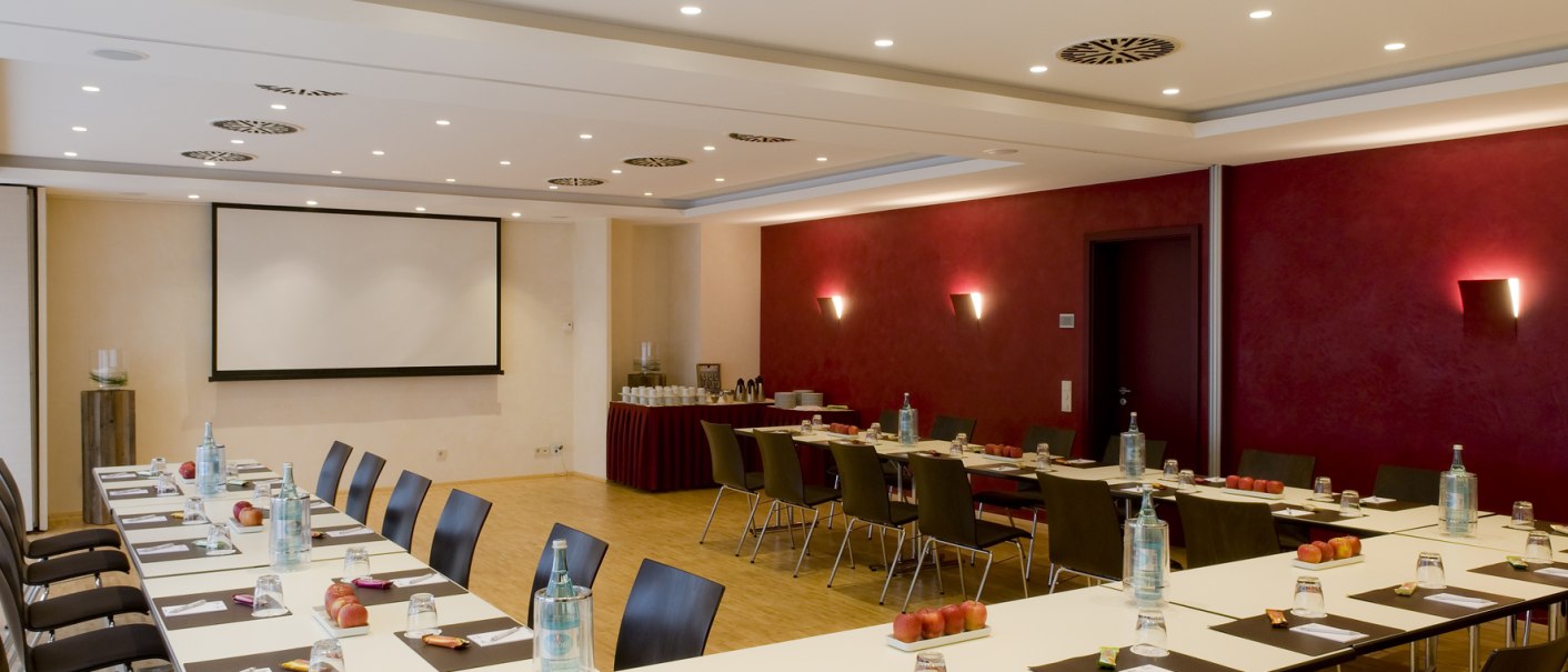 Moderner Tagungsraum mit U-f&ouml;rmiger Tischanordnung, Wasserflaschen, Gl&auml;sern und Notizbl&ouml;cken. Eine Leinwand an der Wand, rote Akzentwand im Hintergrund., &copy; Hotel Gloria