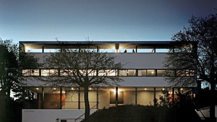 Modernes Gebäude der Weissenhofsiedlung bei Dämmerung, beleuchtet. Klare Linien und große Fenster prägen die Architektur., © Brigida Gonzalez Modernes Gebäude der Weissenhofsiedlung bei Dämmerung, beleuchtet. Klare Linien und große Fenster prägen die Architektur., © Brigida Gonzalez
