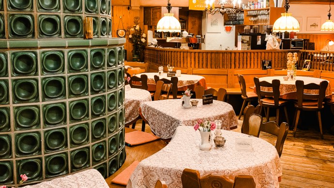Cozy restaurant with wooden furniture, tablecloths and a green tiled stove. Lamps hanging from the ceiling, bar in the background., © Weinstube Kachelofen