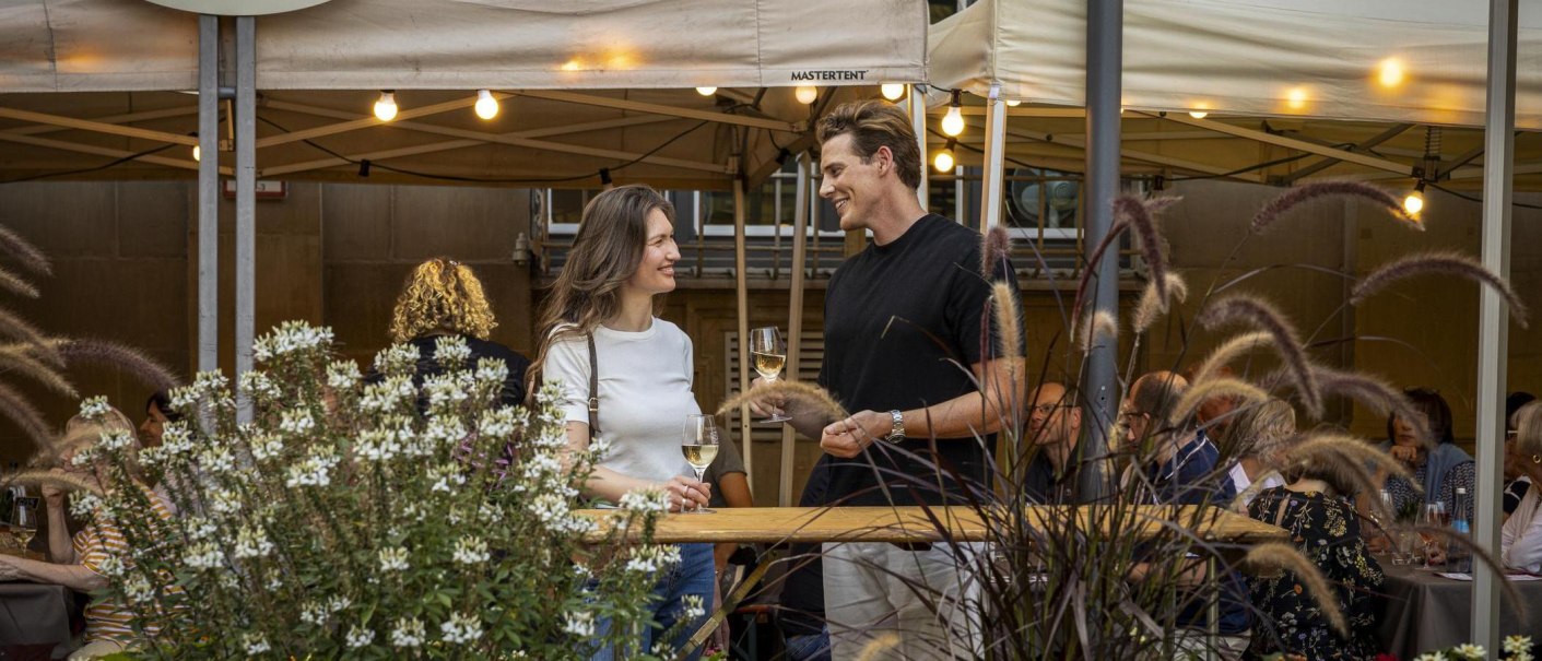 Zwei Personen stehen l&auml;chelnd mit Weingl&auml;sern in der Hand auf dem Stuttgarter Weindorf. Im Hintergrund sind Zelte und weitere G&auml;ste zu sehen., &copy; Stuttgart-Marketing GmbH, Sarah Schmid