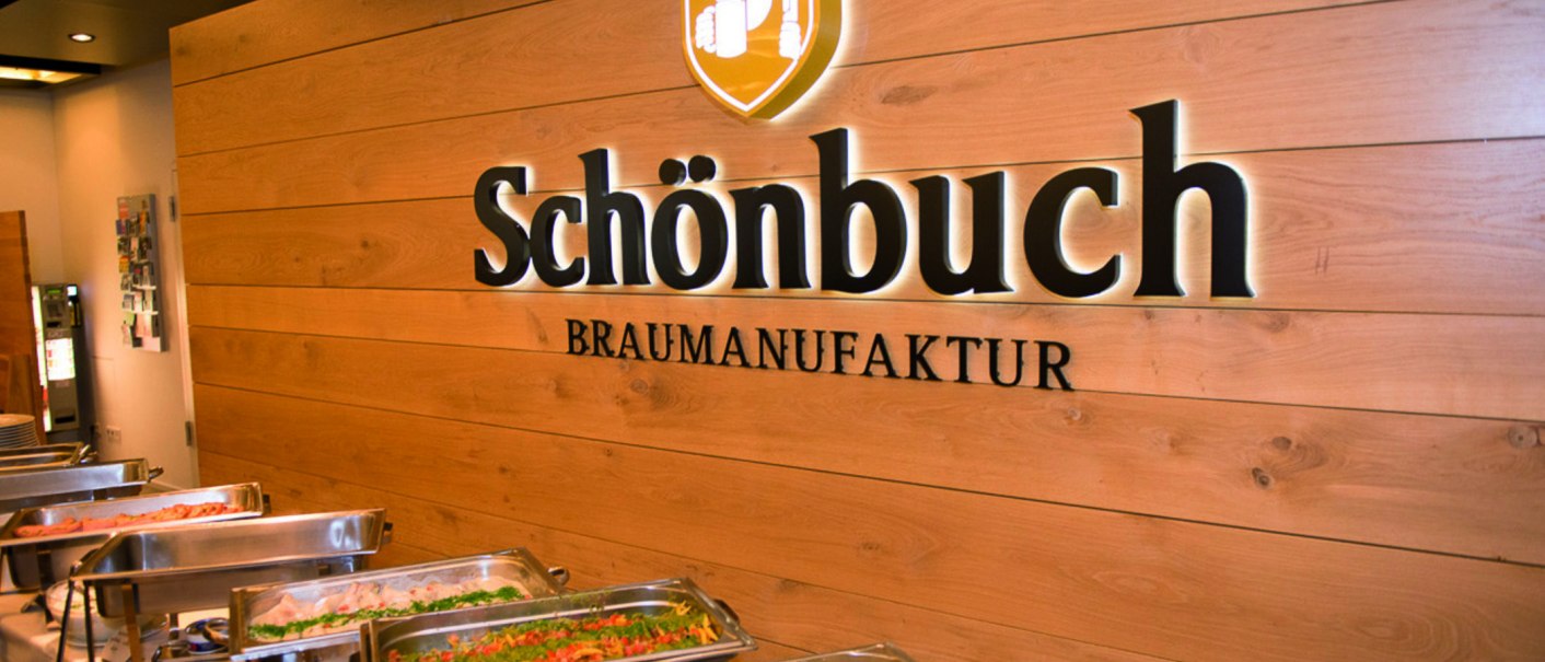 Interior view of a brewery with buffet. "Schönbuch Braumanufaktur" is written on the wooden wall. In the foreground are dishes in warming containers., © © Schönbuchbrauerei, Sebastian Berger
