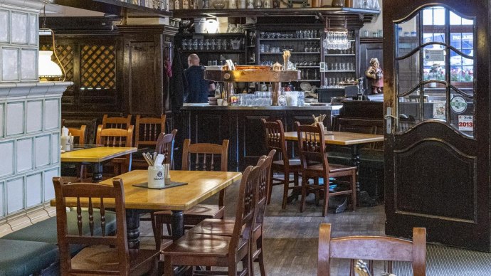 Cozy interior of a restaurant with wooden tables and chairs, a bar in the background and a large tiled stove on the left., © SMG Stuttgart Marketing GmbH - Sarah Schmid