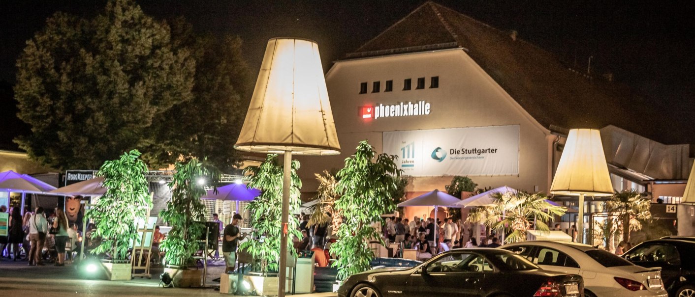 Die Phoenixhalle im R&ouml;merkastell bei Nacht, mit beleuchtetem Au&szlig;enbereich, Pflanzen und geparkten Autos. Menschen genie&szlig;en die Atmosph&auml;re., &copy; Julian Rettig
