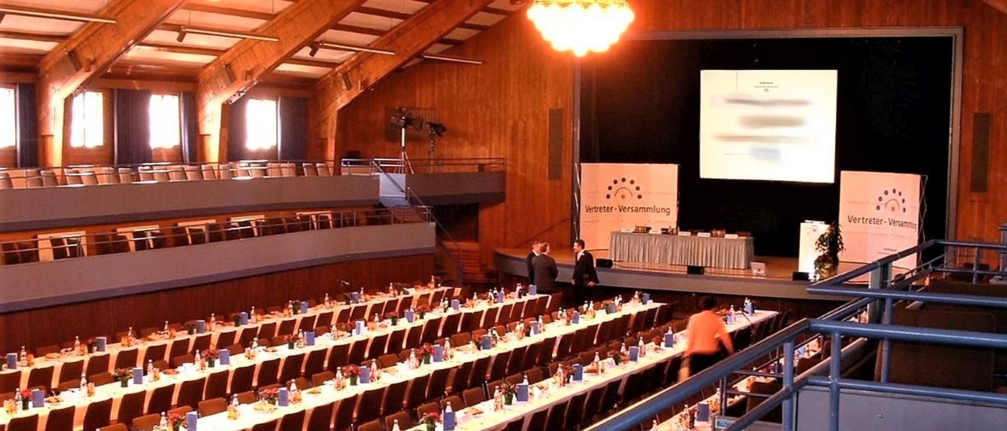Großer Saal der Stadthalle Herrenberg mit langen, gedeckten Tischen und einer Bühne für eine Versammlung. Holzverkleidung und große Deckenleuchten., © Stadt Herrenberg
