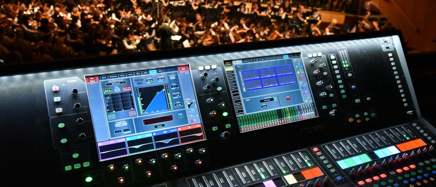 A mixing console with illuminated displays in the foreground, behind it an orchestra and choir on a stage in a concert hall., © Guenter E. Bergmann - Photography A mixing console with illuminated displays in the foreground, behind it an orchestra and choir on a stage in a concert hall., © Guenter E. Bergmann - Photography