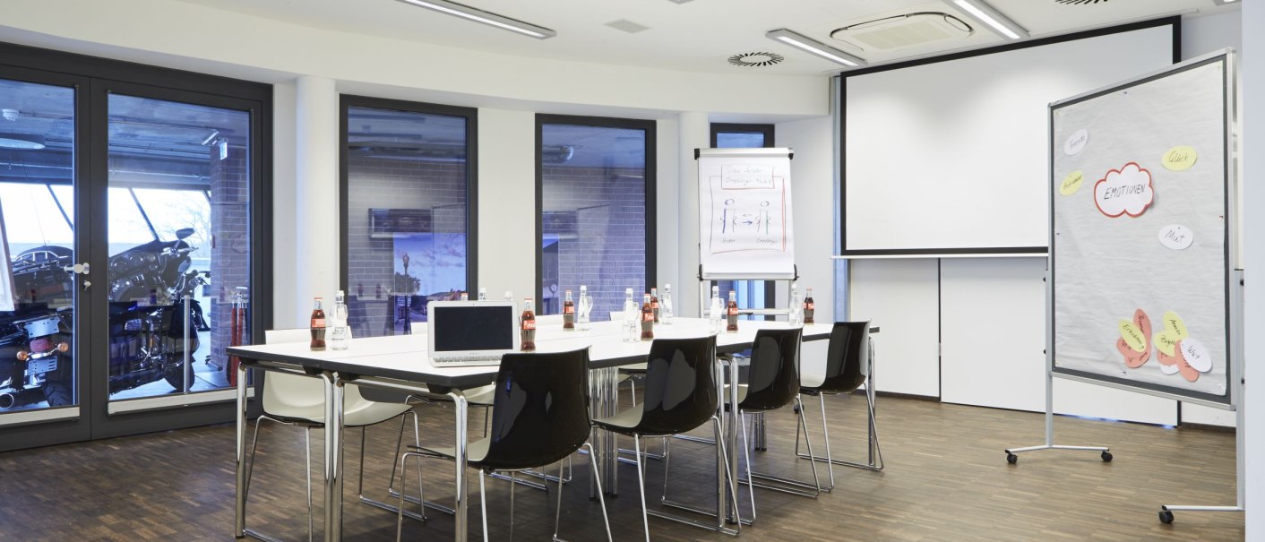Moderner Konferenzraum mit Tisch, Stühlen, Laptop und Flipchart. An der Wand sind große Fenster und ein Projektor ist an der Decke montiert., © MW Region Stuttgart