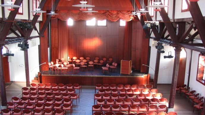 An old gymnasium with wooden beams, a stage and many rows of chairs. The stage is equipped with curtains and a lectern., &copy; Stadt Herrenberg