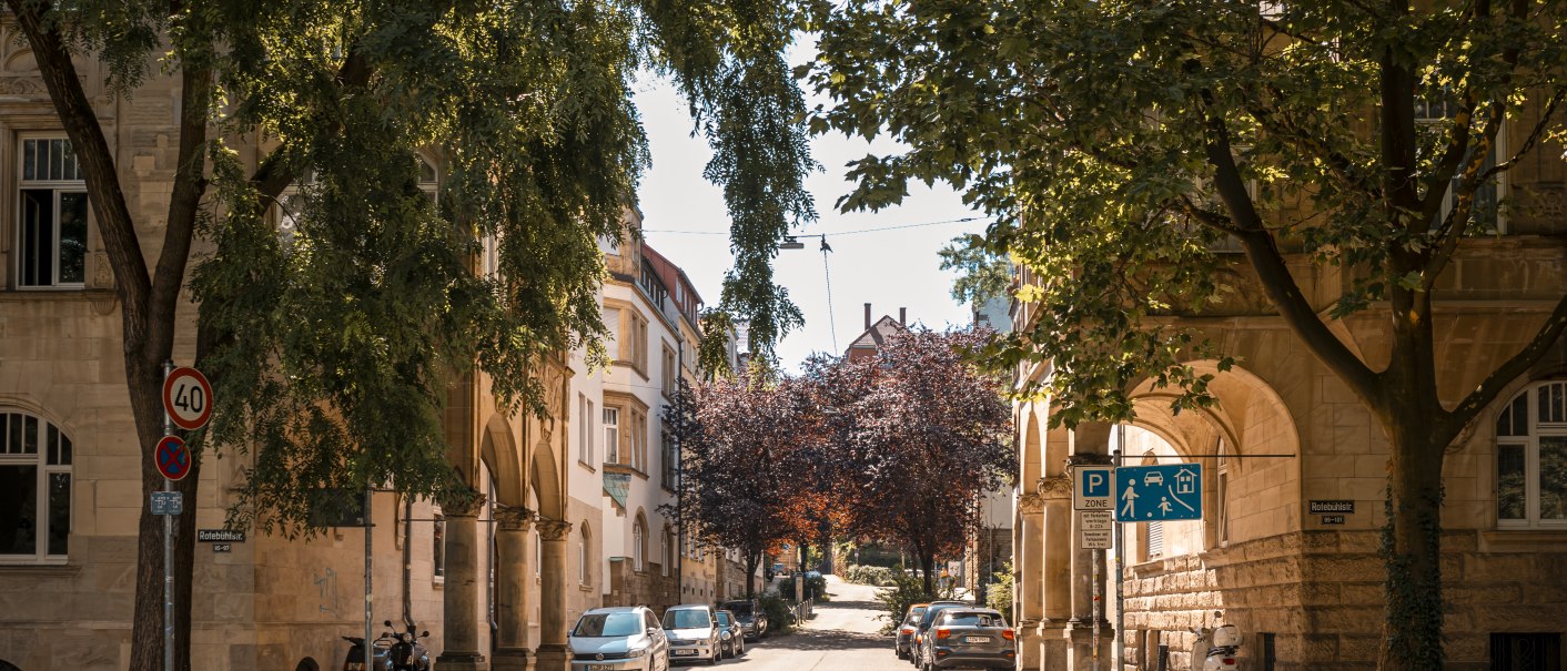 Sonnige Straßenszene mit Bäumen, geparkten Autos und historischen Gebäuden. Verkehrsschilder und ein Gehweg sind sichtbar., © SMG Stuttgart Marketing GmbH - Sarah Schmid