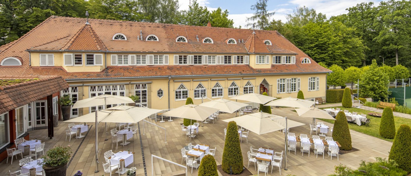 The terrace of the Waldhotel Stuttgart features covered tables with parasols, surrounded by well-tended hedges and trees, under a blue sky., © WHS The terrace of the Waldhotel Stuttgart features covered tables with parasols, surrounded by well-tended hedges and trees, under a blue sky., © WHS