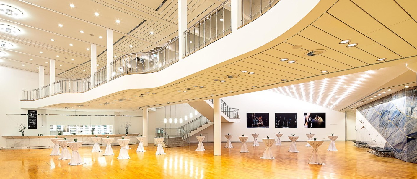 Großes, modernes Theaterfoyer mit Holzboden, Stehtischen und eleganter Beleuchtung. Eine Treppe führt zu einer Galerieebene., © Stauch