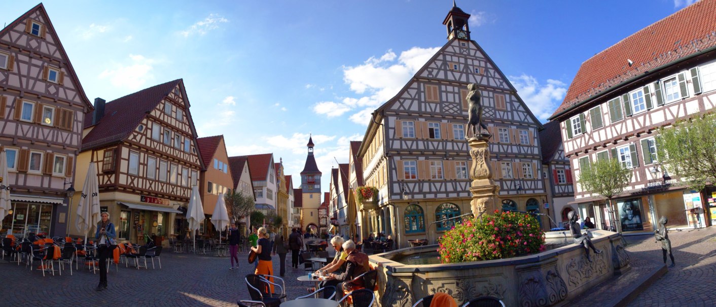 Fachwerkhäuser in Winnenden mit einem Brunnen im Vordergrund. Menschen sitzen und spazieren auf dem gepflasterten Platz unter blauem Himmel., © SMG_Achim Mende