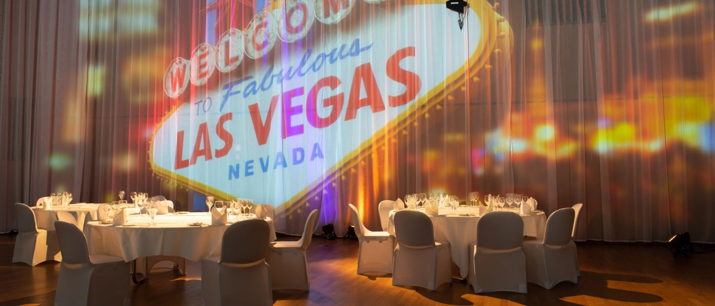 Veranstaltungsraum mit runden, gedeckten Tischen. An der Wand ist das berühmte Las Vegas Schild projiziert. Elegante Atmosphäre mit gedämpftem Licht., © Markus Schwarz Fotograf