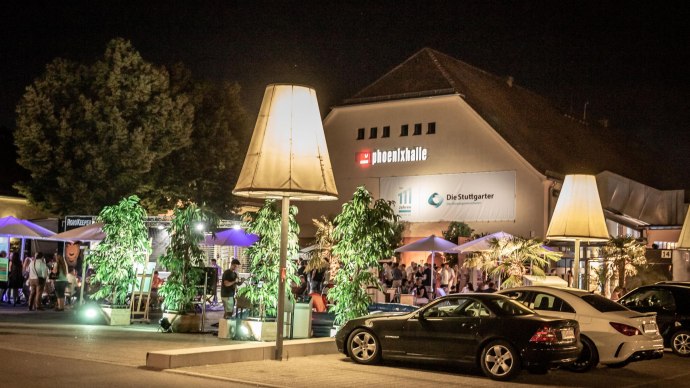 Die Phoenixhalle im R&ouml;merkastell bei Nacht, mit beleuchtetem Au&szlig;enbereich, Pflanzen und geparkten Autos. Menschen genie&szlig;en die Atmosph&auml;re., &copy; Julian Rettig