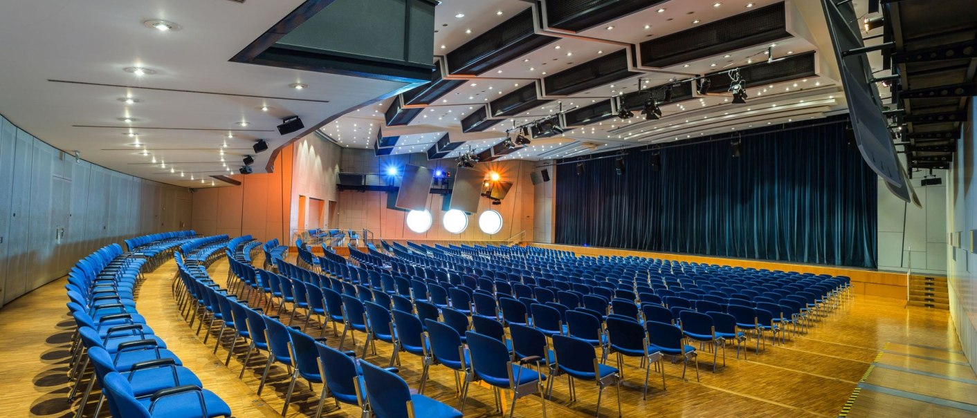 Modern concert hall with rows of blue chairs, wooden floor and a stage with a curtain. The ceiling is equipped with lighting and acoustic elements., © Filharmonie_gf