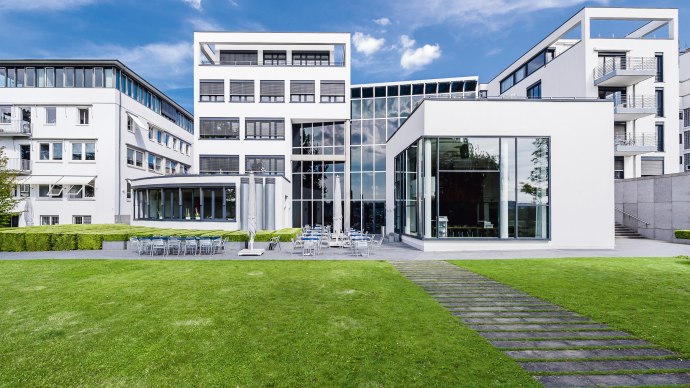 Modernes Geb&auml;ude mit Glasfassade und Rasenfl&auml;che, umgeben von weiteren wei&szlig;en Geb&auml;uden. Klare Linien und gro&szlig;e Fenster pr&auml;gen die Architektur., &copy; Francis Koenig