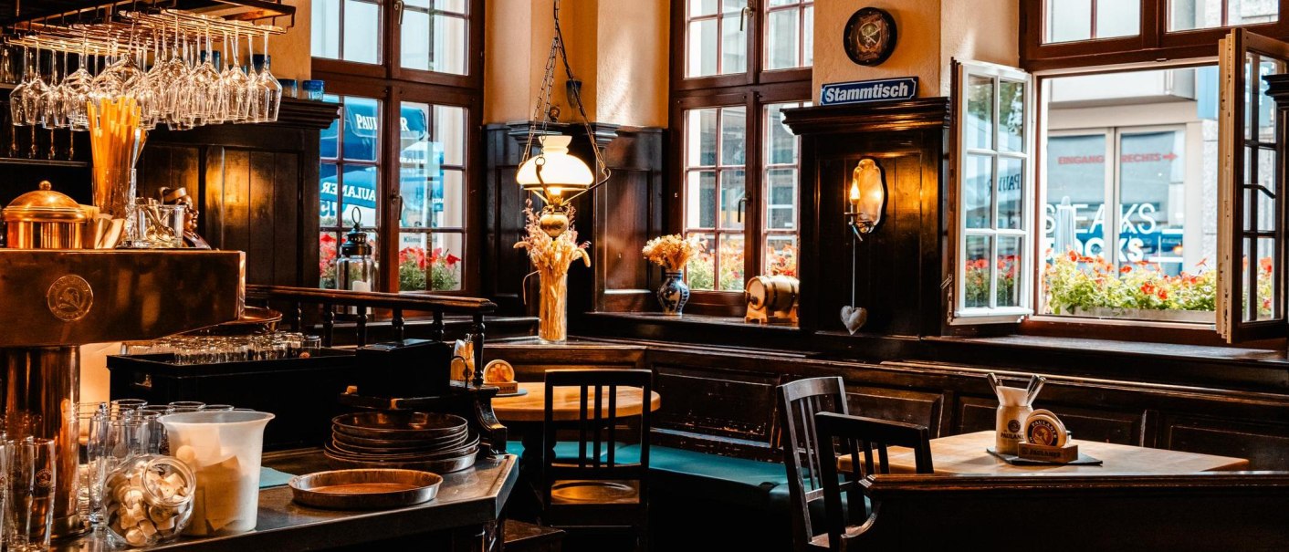 Gemütlicher Innenraum eines Paulaner-Restaurants mit Holzmöbeln, Bar, Gläsern und Dekorationen. Offene Fenster mit Blick nach draußen., © Stuttgart Marketing GmbH, Sarah Schmid