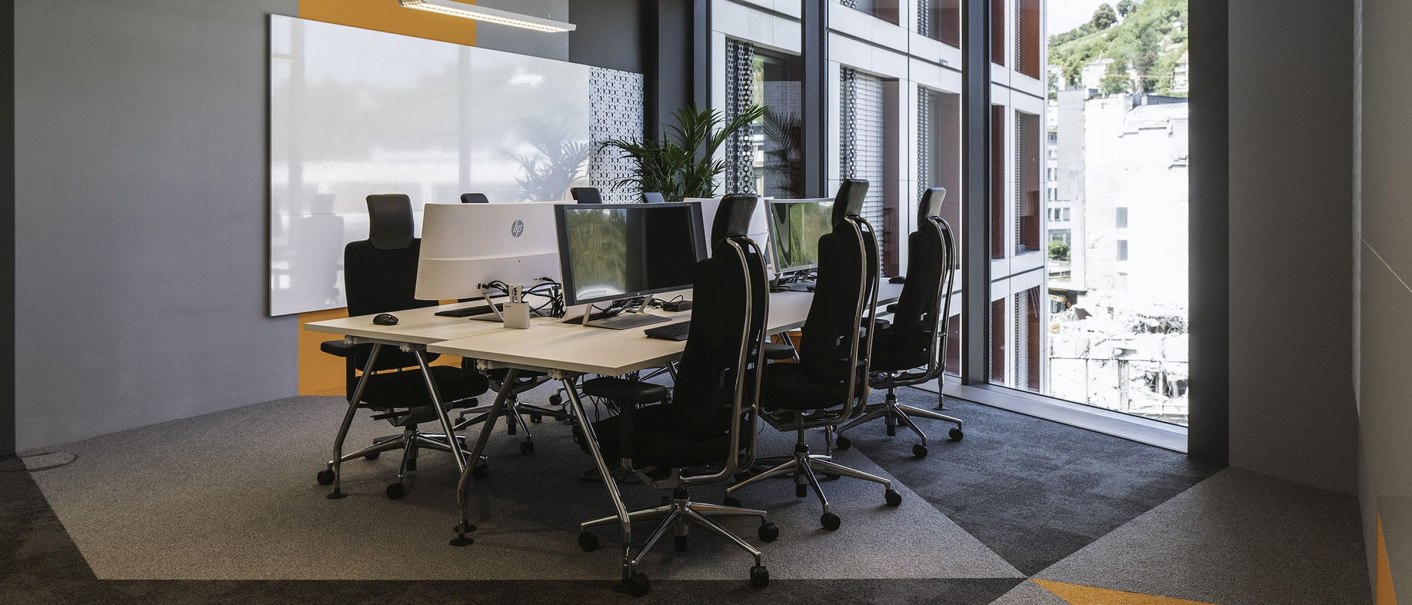 Modern office space with several desks and computers, large windows and geometric carpet pattern. Bright and open atmosphere., © INFOMOTION