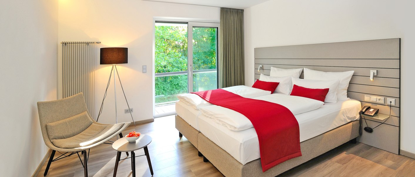 Modern hotel room with double bed, red bedspread, armchair, table and floor lamp. Large windows offer a view of the greenery., © Schlosshotel Monrepos, Hotelzimmer Modern hotel room with double bed, red bedspread, armchair, table and floor lamp. Large windows offer a view of the greenery., © Schlosshotel Monrepos, Hotelzimmer