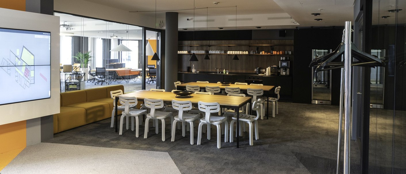 Modern lounge with a large table, white chairs and a kitchenette. Sofas and plants can be seen in the background., © INFOMOTION