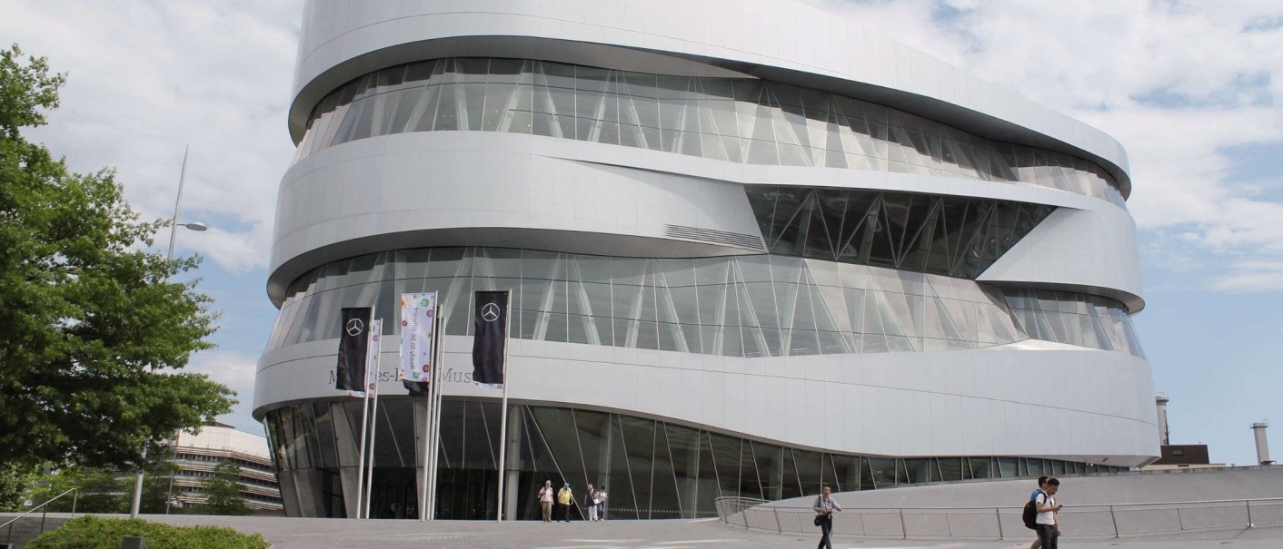 The Mercedes-Benz Museum in Stuttgart shows off its modern, curved architecture with a glass façade and several flags in front of it., © Stuttgart-Marketing GmbH