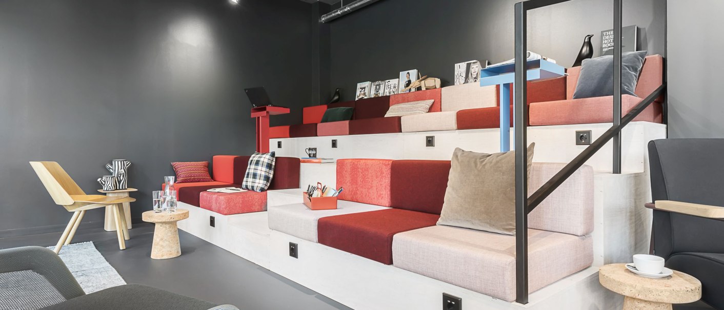 Moderne Coworking-Lobby mit gestuften Sitzbereichen, bunten Kissen und kleinen Tischen auf grauem Boden., © Copysign