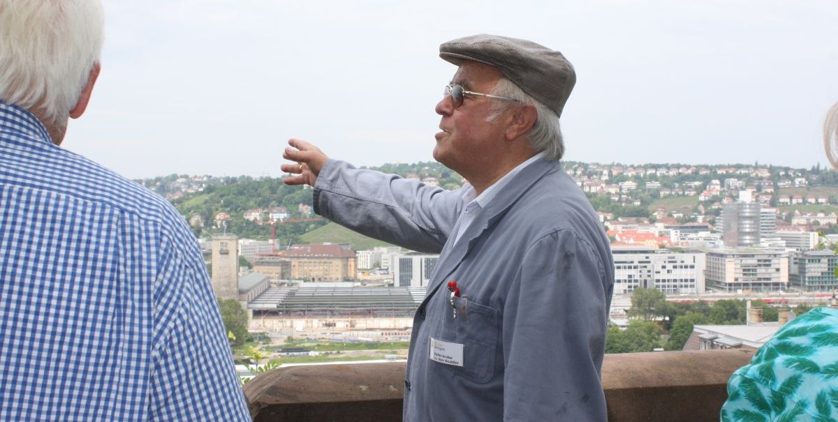 Ein älterer Mann in Arbeitskleidung und Mütze zeigt auf eine Stadtlandschaft. Im Hintergrund sind Gebäude und Hügel zu sehen., © Stuttgart-Marketing GmbH