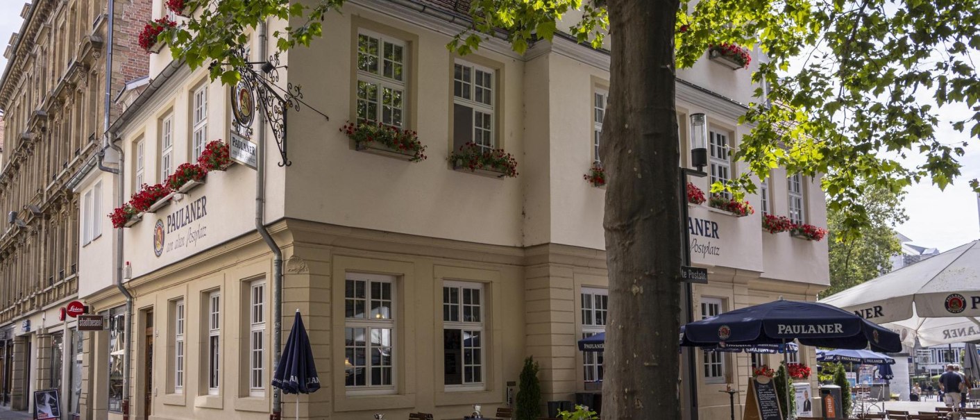 Corner building with Paulaner restaurant, decorated with red flowers. There are parasols and tables in front of the building. Trees provide shade., © SMG Stuttgart Marketing GmbH - Sarah Schmid