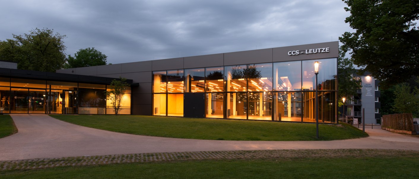 Der CCS Leutze-Saal bei Dämmerung, mit großen Glasfronten und warmem Licht, umgeben von Rasen und Bäumen., © Congress-Centrum Stadtgarten Schwäbisch Gmünd