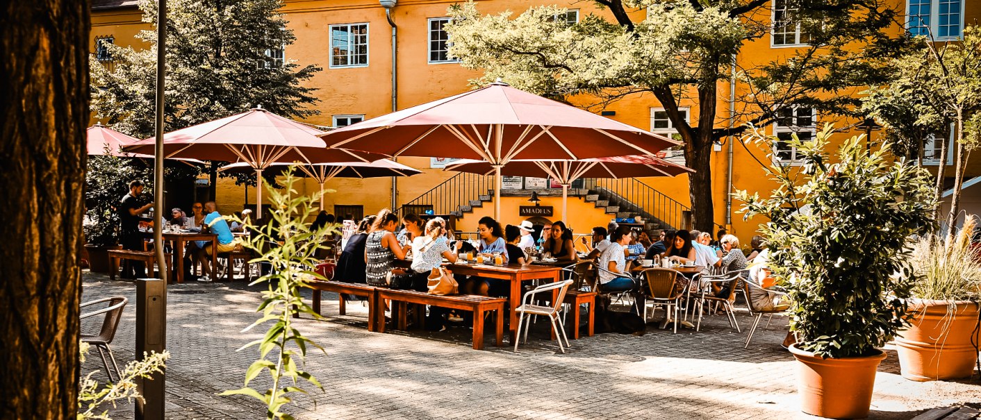 People sit under red parasols in a sunny outdoor area in front of a yellow building., © Amadeus