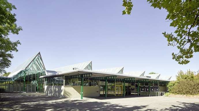 The Glaspalast Sindelfingen with its striking glass roof and green structures, surrounded by trees and a clear sky., © Glaspalast Sindelfingen