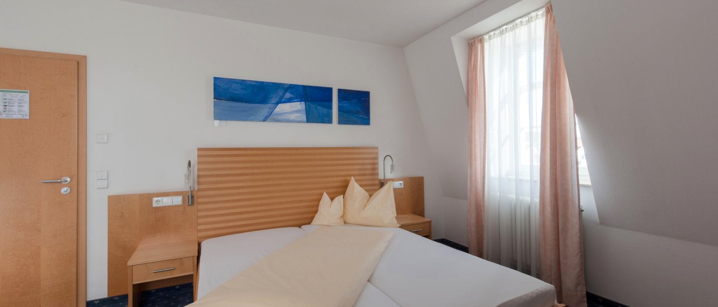 Helles Hotelzimmer mit Doppelbett, Holzmöbeln und blauem Teppichboden. Ein Fenster mit Vorhängen lässt Tageslicht herein., © Landschloss Korntal Helles Hotelzimmer mit Doppelbett, Holzmöbeln und blauem Teppichboden. Ein Fenster mit Vorhängen lässt Tageslicht herein., © Landschloss Korntal