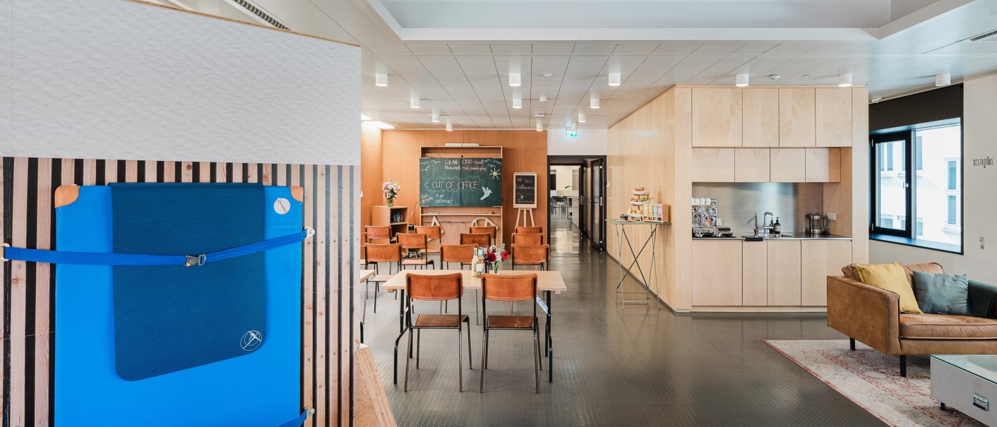 Moderner Büroraum mit Tischen, Stühlen, einer kleinen Küche und einem blauen Sportgerät an der Wand., © OutOfOffice GmbH Moderner Büroraum mit Tischen, Stühlen, einer kleinen Küche und einem blauen Sportgerät an der Wand., © OutOfOffice GmbH