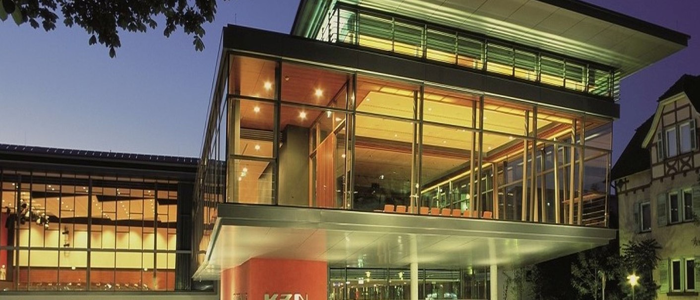 Modern glass fa&ccedil;ade of the K3N building at night, illuminated with warm light. A tree in the foreground, a half-timbered house in the background., &copy; k3nmaisch