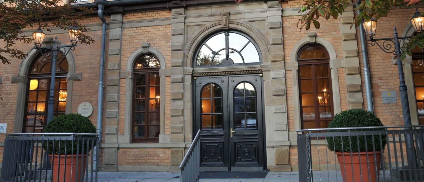 Historisches Gebäude mit großen Fenstern, dekorativen Laternen und Pflanzen in Töpfen. Backsteinfassade mit eleganten Details., © Musikhalle Ludwigsburg