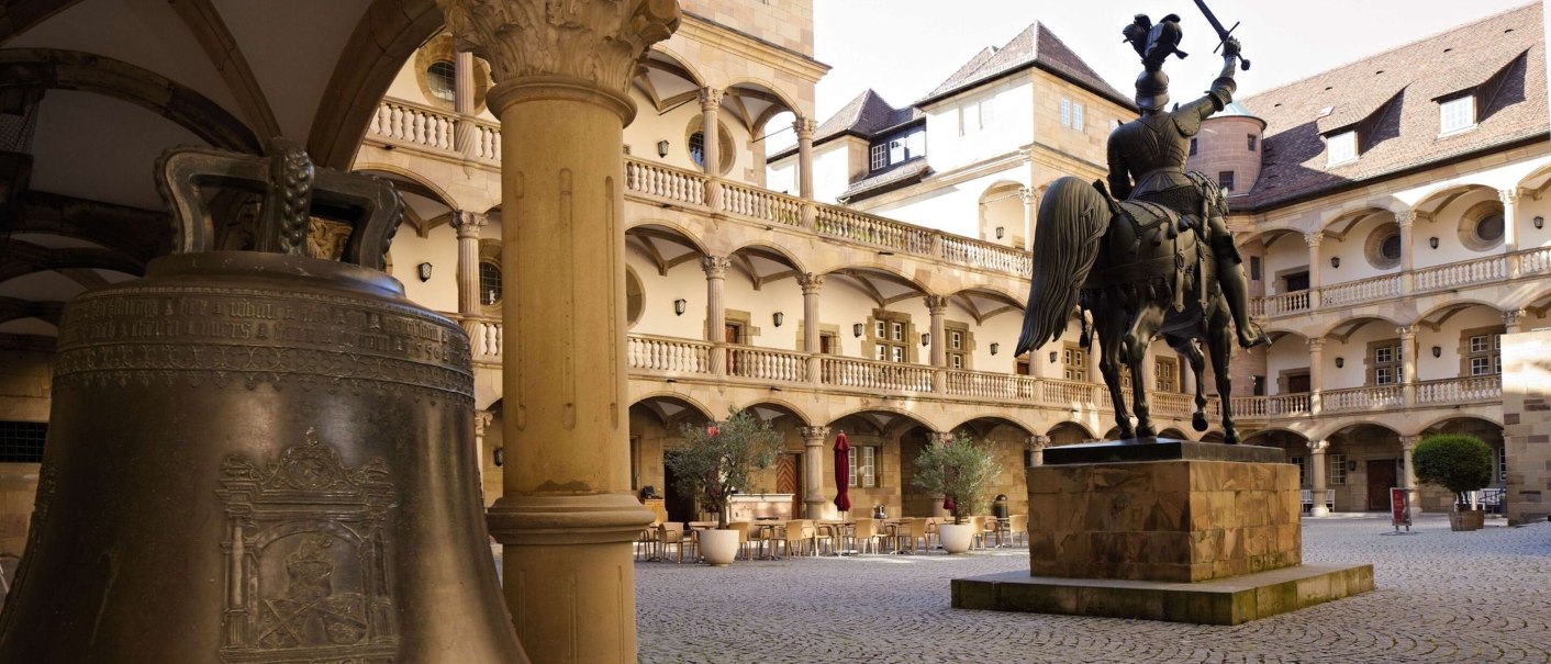 Innenhof eines alten Schlosses mit einer Reiterstatue und einer großen Glocke im Vordergrund. Arkaden und Balkone umgeben den gepflasterten Platz., © Stuttgart-Marketing GmbH Christoph Düpper