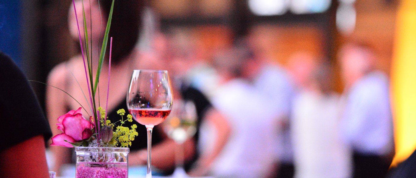 A wine glass and floral decoration on a table, blurred people in the background at an event., © Congress-Centrum Stadtgarten Schwäbisch Gmünd