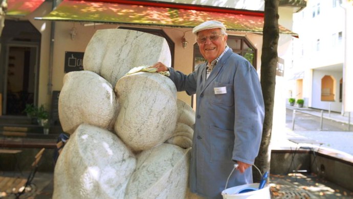 Ein älterer Mann in blauer Arbeitskleidung reinigt eine Steinskulptur. Er trägt eine Mütze und hält einen Eimer in der Hand. Im Hintergrund ist ein Gebäude., © Stuttgart-Marketing GmbH Ein älterer Mann in blauer Arbeitskleidung reinigt eine Steinskulptur. Er trägt eine Mütze und hält einen Eimer in der Hand. Im Hintergrund ist ein Gebäude., © Stuttgart-Marketing GmbH